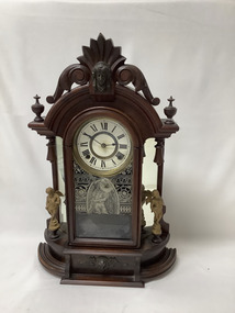 Eight-day mantle clock in an ornate carved wooden case. Plain face with Roman numerals,  under glass. A glass door allows access to the weighted pendulum, and is decocrated with an etching-like transfer showing a bearded Cavalier with a sword. The clock face and pendulum is flanked by a pair of brass or gold coloured cupids, standing on turned wooden pedastels, with mirrors behind them. A carved wooden head is above the face, and another carved wooden head flanked with wings is on the base.