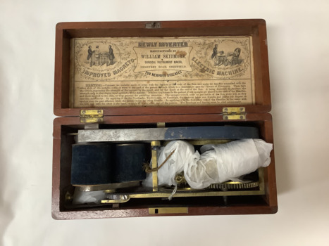 An 1890s magneto shock machine enclosed in a cedar box with instructions on the under-side of the lid. It is actuated by turning the exterior handle which is attached to two gear wheels, one of which has a cord belt that rotates a dual coil.  Metallic wires are attached to two brass handles which when held in each hand conduct the current through the body.
