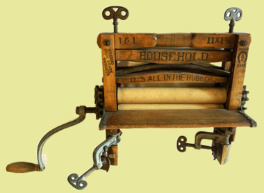 Wooden framed hand wringer with adjustable rubber rollers. Two wing-nuts screw down on a concave steel spring to increase pressure on laundry items. Winding handle engages cogs to turn rollers in the opposite direction. 