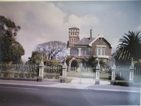 Colour photograph of Maritimo on The Strand, Williamstown. Ornate two storey red brick mansion with square tower on left and tall central chimney. Wrought iron fence with entrance gates to driveway and five pillars.   