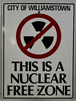 White metal sign, with black and red 'Nuclear free zone' inscription, and the nuclear free emblem in black and white.