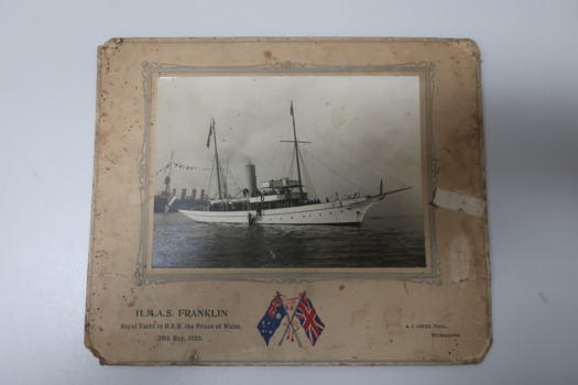 Cardboard framed black and white photograph of the Steam yacht HMAS Franklin, most probably on Port Phillip Bay.  Yacht is under steam. Coloured image of crossed flags of Australia and Britain (symbol of friendship and cooperation) displayed at bottom of frame