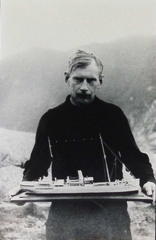The maker of the "Lady Loch" model, holding his model. He was reputed to be a Polish crewman on the ship. He is dressed in a dark jumper, with a view of waves and a rugged coastline in the background. 