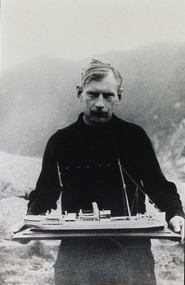 The maker of the "Lady Loch" model, holding his model. He was reputed to be a Polish crewman on the ship. He is dressed in a dark jumper, with a view of waves and a rugged coastline in the background. 