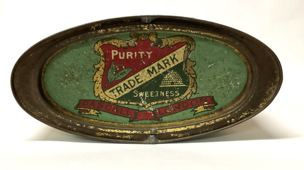 An large oval lidded tin, highly decorated with a gold filigree border, and a large scrolled product name. Main colours are red, pink, green and gold. The price for toffees is shown as 4oz for 3 1/2 d. The lid shows a large shield with a bee hive depicted. 