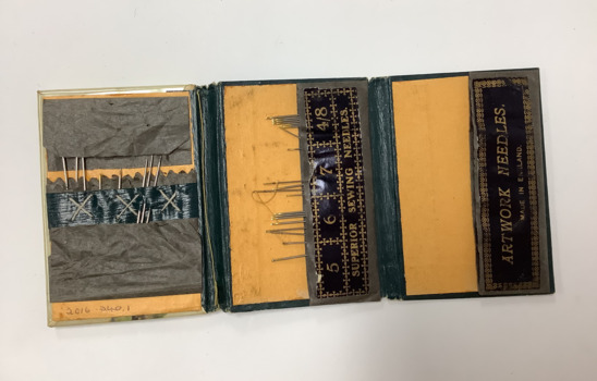 A folding needle case divided into three sections that opens to display a variety of different needles. The cover is blue dyed leather, with a painted portrait of the head of a woman.