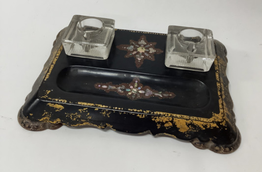 Two glass inkwells on a decorated black wooden base with hollow at the front for pens. Mother of pearl inlay in a star pattern between the ink wells and in pen hollow. Goldleaf decoration around sides of the base.