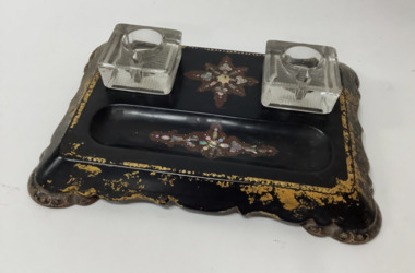 Two glass inkwells on a decorated black wooden base with hollow at the front for pens. Mother of pearl inlay in a star pattern between the ink wells and in pen hollow. Goldleaf decoration around sides of the base.