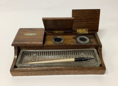 Wooden desk writing set with three inkwells, labelled 'Copying', 'Red' and 'Black' on separate hinged lids. One of the small glass inkwells is missing. A heavy glass decorated pen tray sits in a recess across the front. The wood is prominently grained especially the lids. 