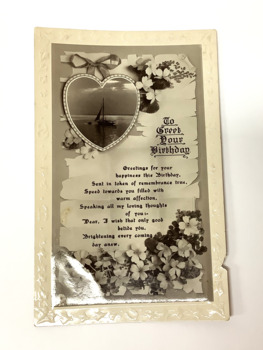 Embossed birthday card (circa 1917) on photographic paper. Decorated with flowers in the upper and lower left, and a bow and heart shape containing a small sailing yacht on the sea. 