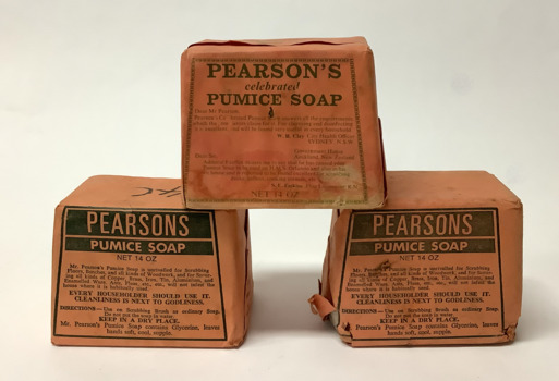 Three large rhomboid shaped cakes of pumice soap, wrapped in original orange paper packaging. Two packages with black text and one package with dark green text.
