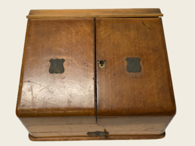 Wooden vintage writing case. Two slanted top opening doors; keyhole on R/H door damaged (no key). Metal badge on each door. Day/date/month display operated by bakelite knobs above display. Four tier holders for stationery/letters etc. Storage for writing implements and pen nibs. 