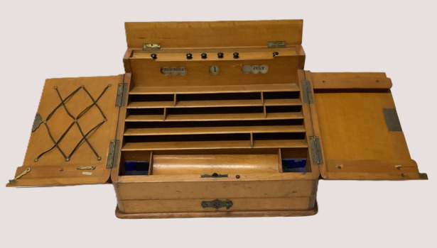 Wooden vintage writing case. Two slanted top opening doors; keyhole on R/H door damaged (no key). Metal badge on each door. Day/date/month display operated by bakelite knobs above display. Four tier holders for stationery/letters etc. Storage for writing implements and pen nibs. 