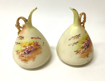 Pair of small urn shaped china jugs, with ornate gold rimmed handles, hand painted with sprays of yellow and mauve flowers. Very small pouring holes and spout. Probably used for hair oil, and as a dressing table accessory.