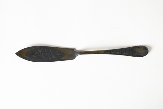 A tarnished dark brown and grey metal butter knife with engraved and embossed details on the knife and handle