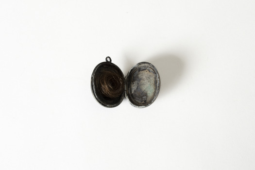 An open locket, the left hand side has a coil of reddish human hair in it, the other appears to be a degraded and illegible photo of a person. 