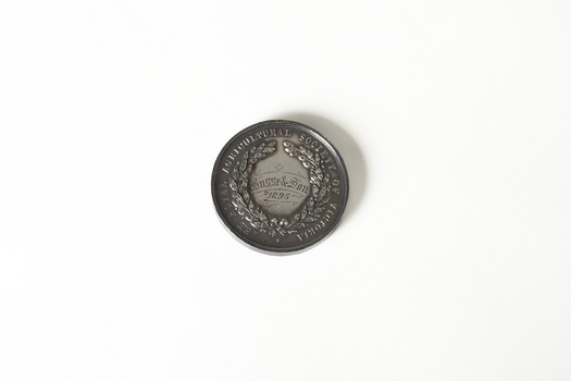 Front side of a small, round, dark-coloured medal depicting an agricultural country scene in the background. In the foreground there is a woman in a toga holding up a wreath. 