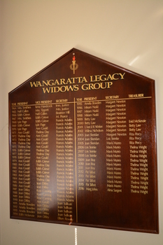 Stained brown timber five sided with Legacy crest and list of names in gold coloured paint.