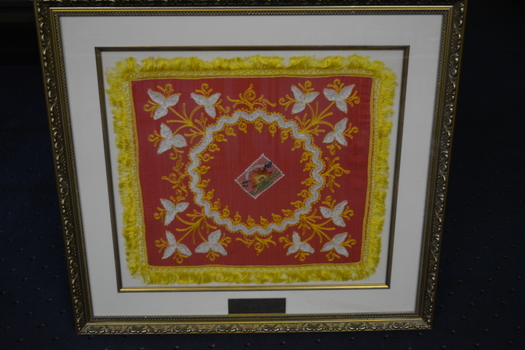 Ornate gold coloured plastic frame containing a pink cloth square embossed in yellow and white thread and edged in yellow fringe.