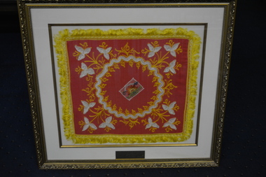 Ornate gold coloured plastic frame containing a pink cloth square embossed in yellow and white thread and edged in yellow fringe.