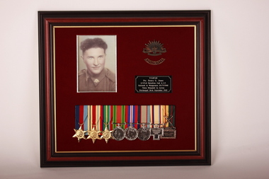 Brown timber frame with maroon velvet mount  containing 8 medals, black and white photograph, two brass badges and a black plaque with white inscription .