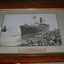 Carved timber frame containing sepia coloured photograph of ship being towed by tug boat from wharf crowded with people 