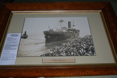 Carved timber frame containing sepia coloured photograph of ship being towed by tug boat from wharf crowded with people 
