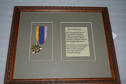 Timber look frame containing medal on yellow/blue/red ribbon and paper with black writing on grey cardboard surround.