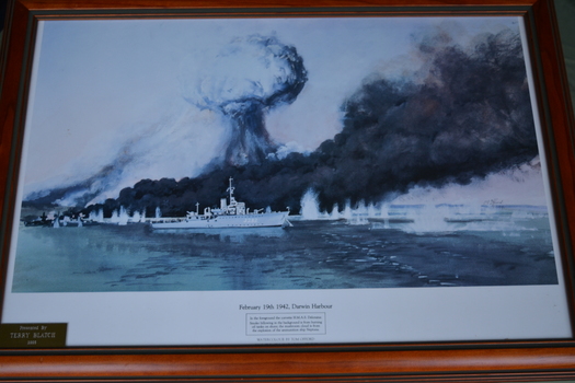 Brown timber frame containing blue black and white print of ships being bombed.