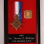 Black timber frame containing bronze coloured star shaped metal medal with red blue and white striped ribbon and a gold-coloured metal badge/medallion.