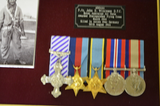 Brown timber frame containing 5 metal medals, photograph of Airman, flying patch and plague on red felt background