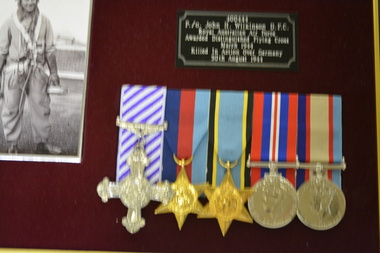 Brown timber frame containing 5 metal medals, photograph of Airman, flying patch and plague on red felt background