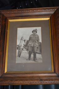Ornate dark timber frame housing photograph of soldier wearing pith style helmet with plum standing in front of large two wheel cart/dray