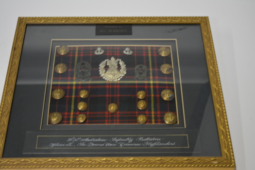 Gold coloured metal frame with green mount containing 3 silver and 2 green badges 15 gold coloured buttons on tartan cloth background. 