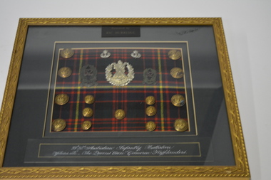Gold coloured metal frame with green mount containing 3 silver and 2 green badges 15 gold coloured buttons on tartan cloth background. 