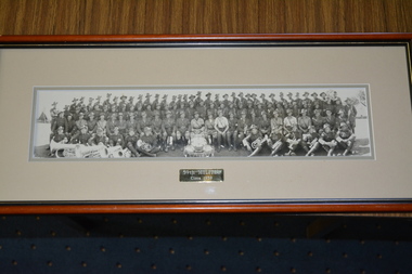 Brown timber frame containing cream coloured mount and photograph of large group of soldiers and bandsmen