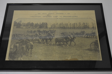 Black plastic frame with black mount with newspaper article depicting mounted soldiers and horse driven cannons.