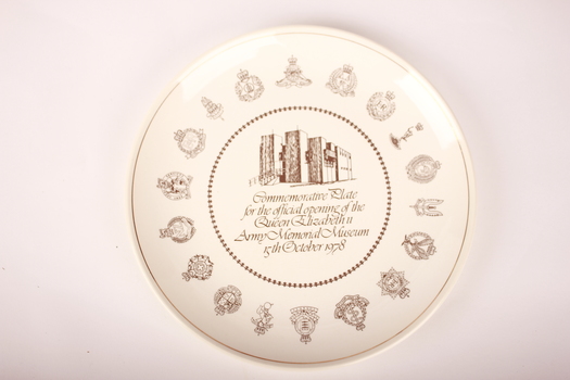 Round white ceramic plate with various royal, imperial and military gold emblems bordering an image of a brown building.