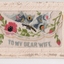 White rectangular postcard with embroidered pink flowers, green foliate and blue winged butterfly decoration on front.  