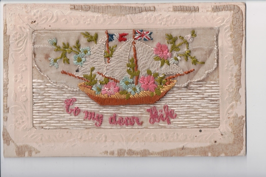 White rectangular postcard with embroidered sailing boat with flying french flag and union jack.  Also pink, blue flowers and green foliate decoration on front. 