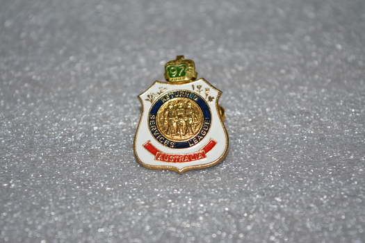 Gold badge with white on outside, crown on top, blue circle in the middle with gold in centre, Has red banner at bottom.