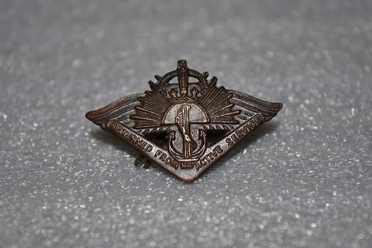 Dark brown metal badge with boomerang at bottom, anchor above, rising sun in middle and crown at the top.