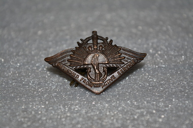 Dark brown metal badge with boomerang at bottom, anchor above, rising sun in middle and crown at the top.