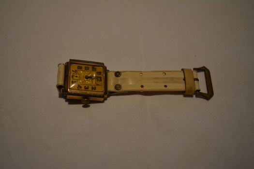 Gold coloured metal watch with square face and white plastic watch band.
