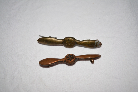 Two brass coloured metal badges in shape of a propeller - one badge slightly larger appears handmade from silver metal and has rear clips attached.