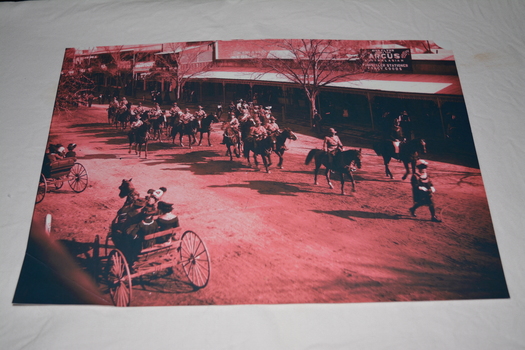 Black white and dark pink photograph of soldiers on horseback being lead by Town Crier passing horse and buggies along main street.