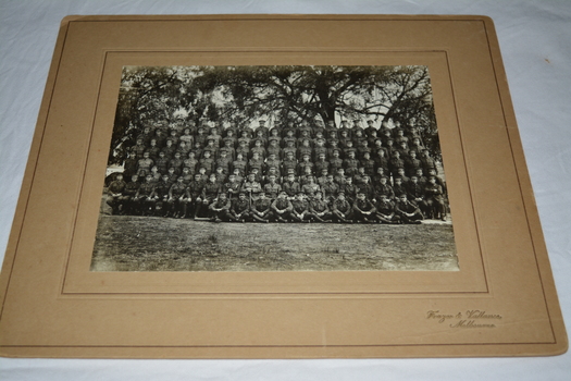 Black and white photograph mounted on light brown cardboard of large group of soldiers standing and sitting in seven rows taken outdoors under trees.