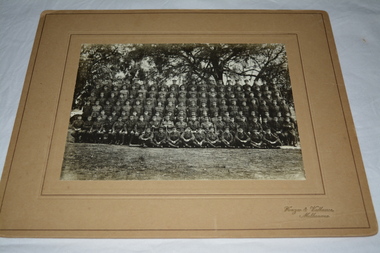 Black and white photograph mounted on light brown cardboard of large group of soldiers standing and sitting in seven rows taken outdoors under trees.