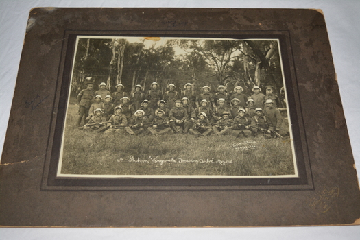 Black and white photograph mounted on dark grey cardboard depicting three rows of soldiers wearing bush hats and front row holding rifles