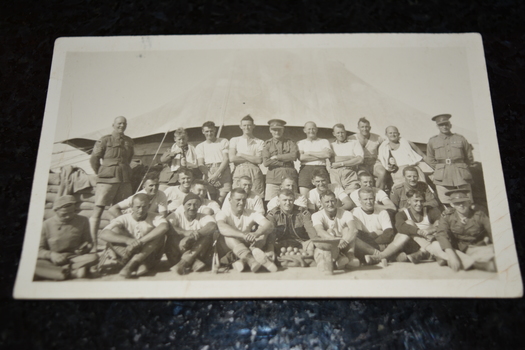 Black and white photograph of men in three rows some in uniform others in white tops in front of large tent.